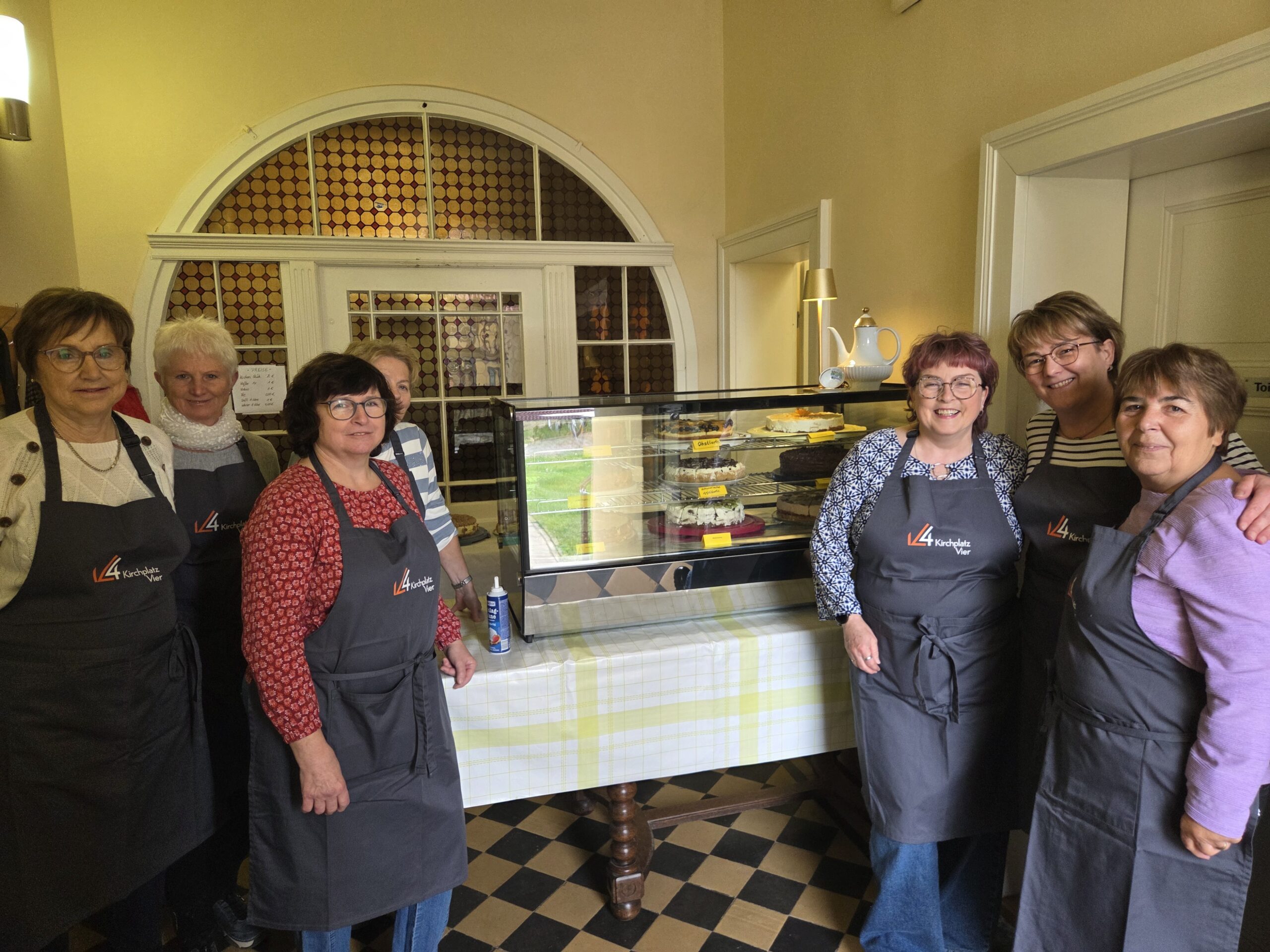 Sechs Frauen in Schürzen stehen lächelnd neben einer Auslage mit Kuchen in einem gemütlichen Innenraum mit karierten Bodenfliesen und einer gläsernen Kuchentheke.
