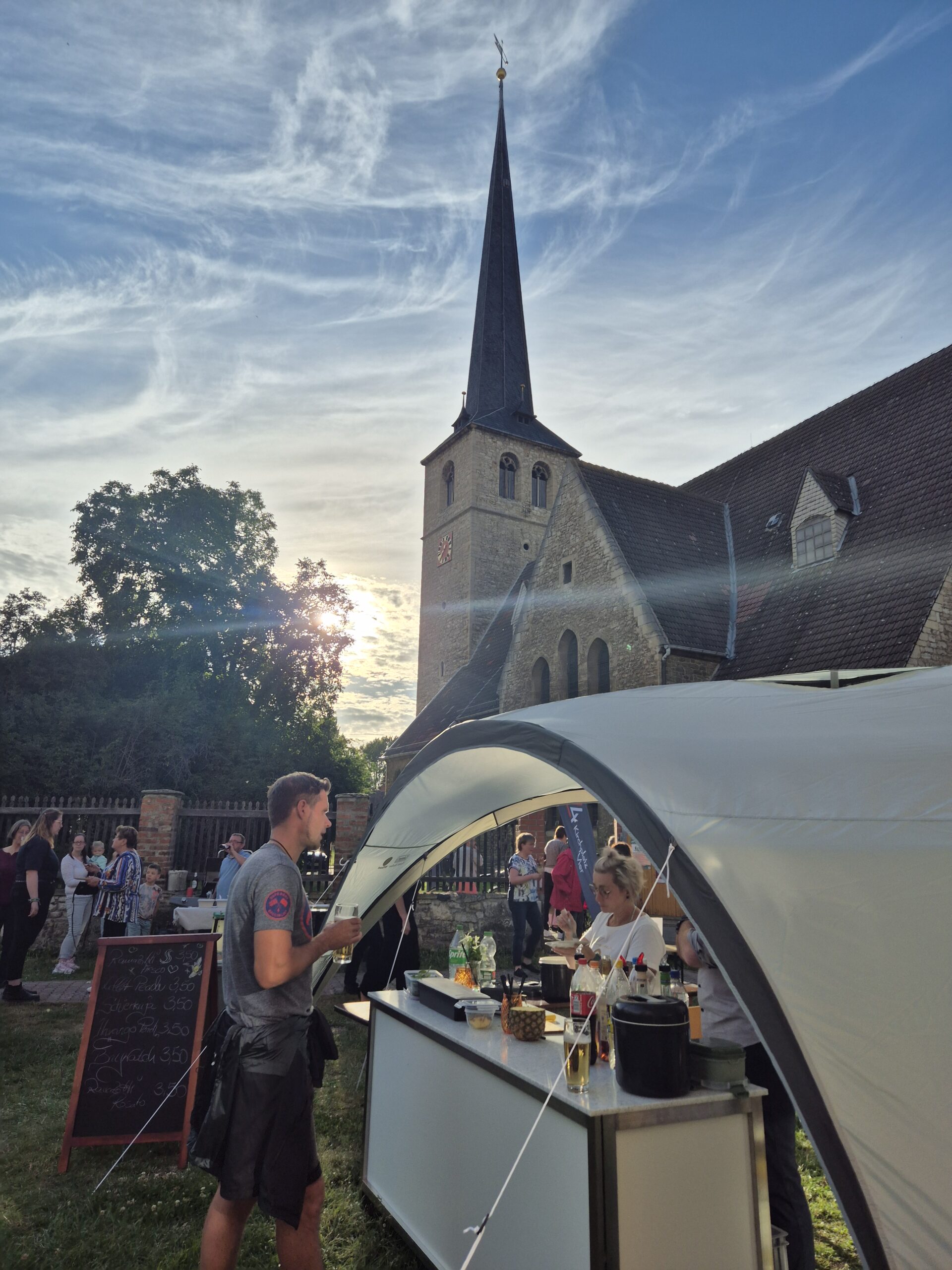Bei Sonnenuntergang versammeln sich Menschen an einem Imbissstand im Freien in der Nähe einer historischen Kirche mit hohem Kirchturm; im Vordergrund ist die Speisekarte auf einer Kreidetafel zu sehen.